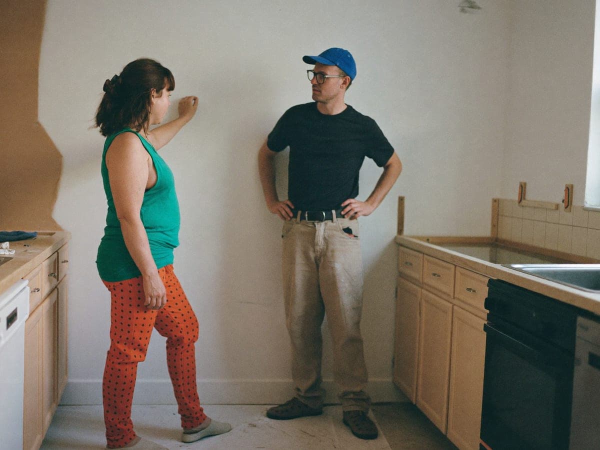 A homeowner in a half-finished kitchen pointing at a wall while a tradesperson stands listening.